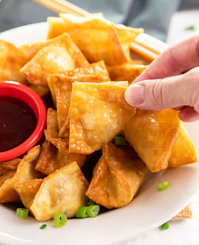 Crispy wontons are baked or fried to golden perfection and filled with an easy cream cheese Crab Rangoon filling. Plus, they're easy to make ahead of time for seamless entertaining! A close up shot of a hand holding fried crab rangoon.