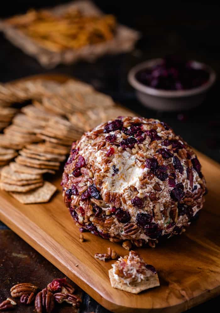 This sweet and savory Pineapple Pecan Cheese Ball has an unbeatable flavor combination that includes pineapple, pecans, bacon, two cheeses, and more! It’s ready in minutes, can be made ahead of time, and can even be baked! | The Cozy Cook | #Appetizer #Cheeseball Cheese ball covered in dried cranberries and pecans on a wooden cutting board with crackers