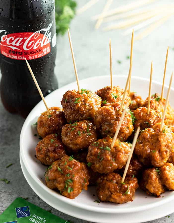 These spicy sausage balls come together with nothing more than cheddar, cream cheese, sausage, and bisquick! Make them ahead of time and keep them warm in the Crock Pot! #christmasappetizers #appetizerideas #fingerfood #newyearseverrecipes White plate filled with spicy sausage bites and a coca-cola zero sugar bottle in background.