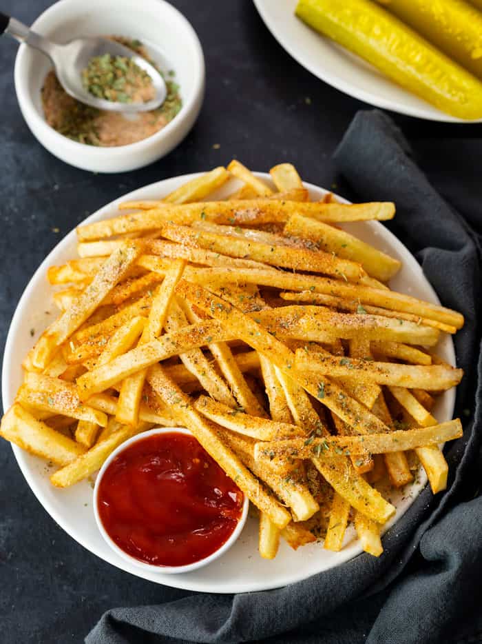 Overhead view of a white plate with a pile of seasoned french fries with ketchup for dipping and a cup of seasoning in the background.