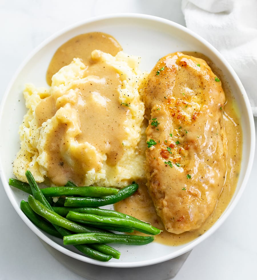 A white plate with chicken and gravy with mashed potatoes and green beans.