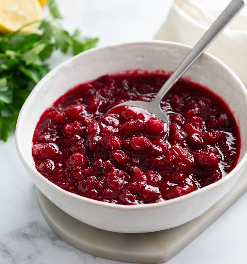 Homemade Cranberry Sauce in a white bowl with a spoon and parsley in the background.