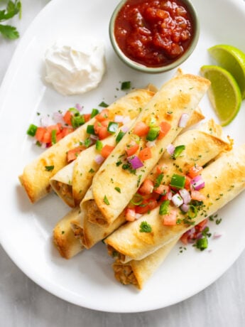 Chicken Taquitos on a white serving platter with salsa, sour cream, lime slices, and toppings.