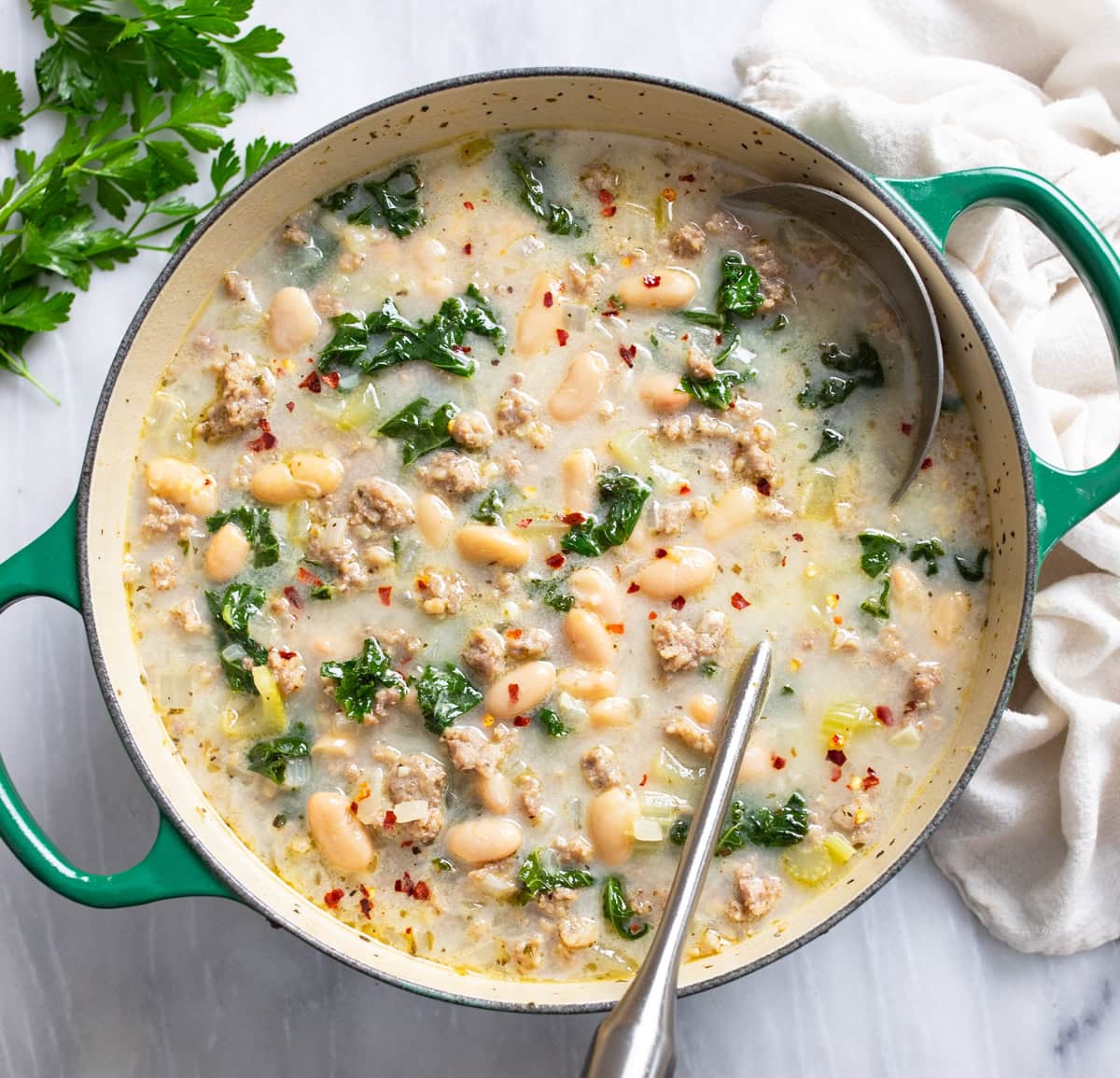 A soup pot filled with Sausage and White Bean soup with kale and red pepper flakes, with a ladle on the side.
