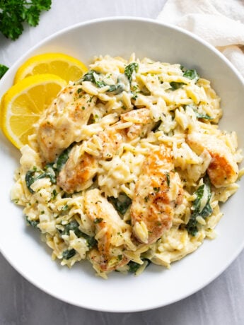 Lemon Chicken Orzo in a white bowl with lemon slices on the side and chicken and spinach in a creamy sauce with orzo.