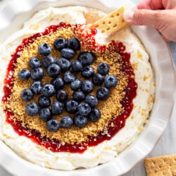 Cheesecake Dip with red jam, buttery graham cracker crumbs, and fresh blueberries with a hand dipping a cookie into it on the side.